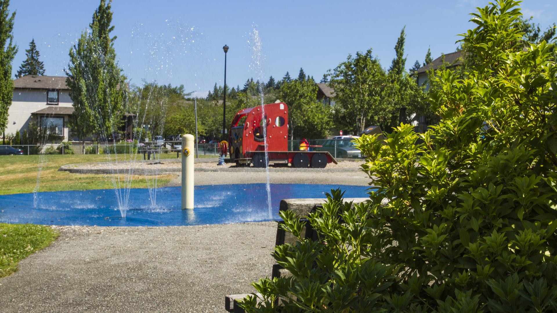 A park bench on the edge of the park, next to a small bush and looking toward the playground.